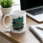 HL NOVA 11oz white ceramic coffee mug with 'Mind Your Own Business' teal and gold design, sitting on a modern wooden office desk next to an open laptop, spiral notebook, and potted plant.
